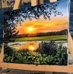 an easel with a painting on it in front of some water and trees at sunset