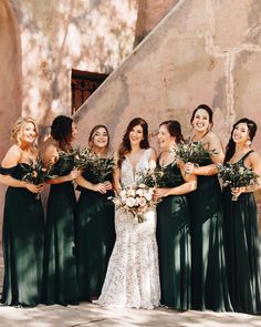 a group of women standing next to each other in front of a stone wall holding bouquets