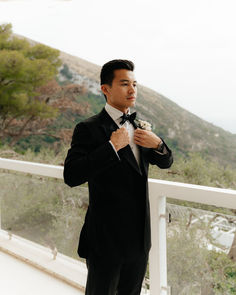 a man in a tuxedo standing on a balcony looking at the camera and adjusting his bow tie