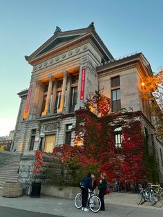 two people standing in front of an old building with red leaves on the trees and bicycles parked outside