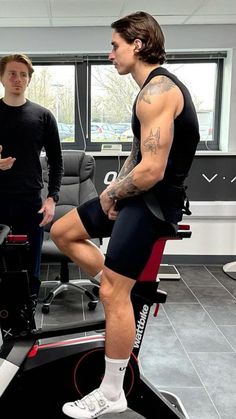 two men standing in an office with one sitting on a stationary exercise bike and the other standing up