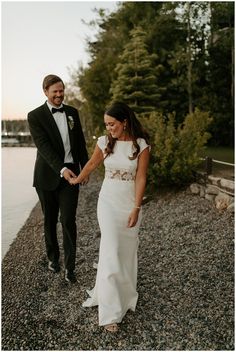 a bride and groom holding hands while walking along the beach at sunset with trees in the background