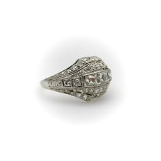 This platinum diamond ring is on the cusp of the Edwardian and Art Deco eras. The centerpiece of the ornate ring are three Peruzzi cut bezel set diamonds —one is an elongated rectangular shape, while the diamonds on either side are truncated, cut specifically for this ring. The larger diamonds are surrounded by a selection of 40 single and Old Mine Cut diamonds that are bead set into the ring. The ring has layers divided by milgrain work that tie together the lace-like patterns of the Edwardian era and the geometric designs of the Art Deco era in alternating bands. There is an added detail of a hand engraved scrolling foliate on each side of the ring. The ring has open work on the bottom that lets in the light and also reveals the triangular bottom shape of the diamonds—a nice touch that h White Art Deco Baguette Cut Jewelry, Art Deco Platinum Diamond Ring With Single Cut Diamonds, Art Deco Oval Platinum Diamond Ring, Formal Diamond Ring With Prong Setting And Rectangle Stone, Luxury Diamond Ring With Rectangular Stone, Timeless Silver Wedding Ring, Square Cut, Luxury Rectangular Platinum Jewelry, Formal Square Cut Diamond Ring With Rose Cut Diamonds, Elegant Square Cut Diamond Ring