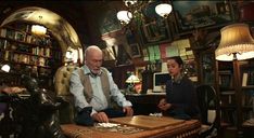 two people sitting at a table in a room with bookshelves and other items