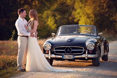 a bride and groom standing in front of an old mercedes benz coupe on the road