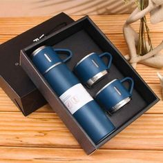 two blue coffee mugs in a black box on a wooden table next to a vase