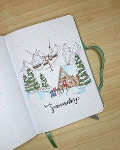 an open notebook with a drawing of a cabin and ski lift in the background on a wooden table