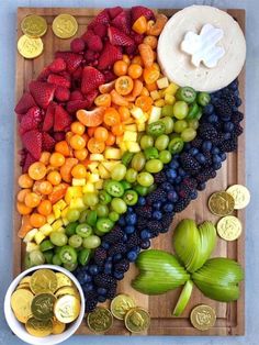 a wooden cutting board topped with lots of fruit next to two bowls of gold coins