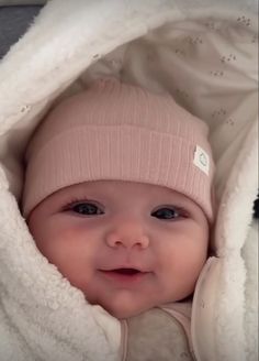 a baby wearing a pink hat under a white blanket and looking at the camera with a smile on its face