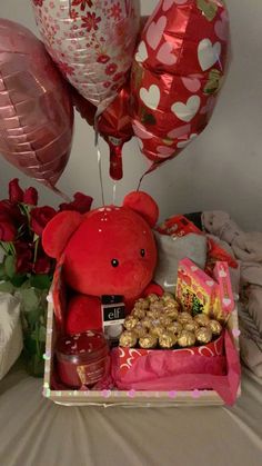 a red teddy bear sitting on top of a box filled with food and balloons in the background