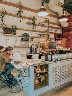 people are sitting at tables in a coffee shop with shelves and hanging lights above them