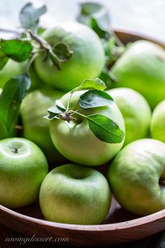 a wooden bowl filled with green apples on top of a table next to some leaves