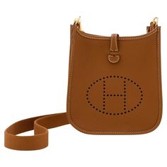 Hermès mini Evelyne in gold Clemence leather complemented with gold-tone hardware and white stitching. It features a perforated "H" design, a removable shoulder strap, an open interior, and a snap lock closure. Date stamp is W 2024 The dimensions are 16cm L x 20cm H x 5cm D. The bag comes complete with a Hermès box, dust bag, removable strap, care book. Hermes Small Crossbody Bag, Hermes Crossbody Bag, Date Stamp, H Design, Snap Lock, Hermes Box, Fashion Handbags, Purses Crossbody, Gold Hardware