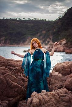 a woman with red hair wearing a blue dress standing on rocks near the water and holding her arms out