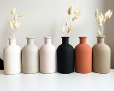 five different colored vases are lined up on the table, one is empty and the other has dry grass in it