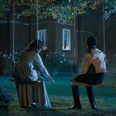 two people sitting on swings in front of a house at night, one holding the other's hand