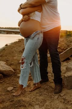 a pregnant woman is hugging her husband while he stands next to the water at sunset