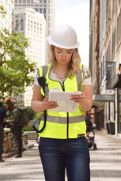 a woman wearing a hard hat and safety vest is looking at a piece of paper