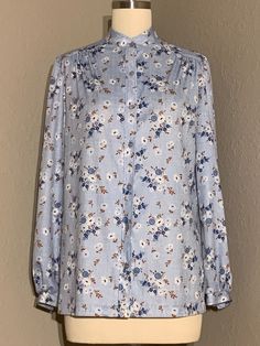 Absolutely darling 1970's button up blouse in a cute country floral print with crosshatch background, shades of blue, white and brown throughout. Blouse has blue pearlescent buttons up the front to a high collarless neckline. Beautiful ruching at the shoulders and long sleeves with single button cuffs. Made of a medium weight polyester fabric. Original brand tag no longer present, labeled a vintage size 11/12. Blouse would best fit a modern women's size medium (please see measurements below for Blue Blouse With Vintage Print For Spring, Classic Floral Print Tops For Daywear, Classic Floral Print Blouse For Fall, Classic Floral Print Tops For Spring, Spring Light Blue Blouse With Button Closure, Light Blue Blouse For Fall Daywear, Classic Floral Print Spring Tops, Classic Printed Tops For Spring, Classic Light Blue Blouse For Spring