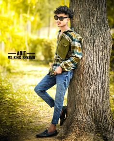 a young man leaning against a tree in the woods wearing sunglasses and a plaid shirt