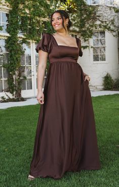 a woman in a long brown dress is standing on the grass and posing for a photo