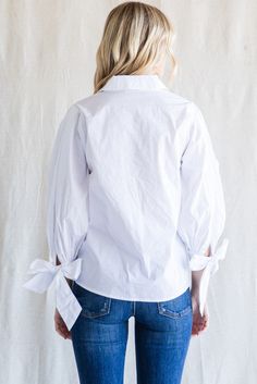 a woman standing in front of a white wall wearing jeans and a white shirt with tied up sleeves