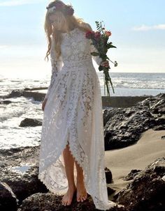 a woman in a white dress standing on rocks near the ocean and holding a bouquet of flowers