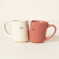 two coffee mugs with eyelashes on them sitting side by side, one pink and the other white