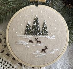 a cross stitch christmas ornament hanging on a tree branch with pine trees in the background