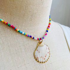 Embrace the essence of summer with this exuberant, colorful beaded necklace, featuring a stunning natural seashell pendant encased in a delicate golden trim. Each bead is meticulously selected to create a kaleidoscope of joy around your neck, reminiscent of a sun-kissed day at the beach or a vibrant festival dance. The heart of this necklace is the seashell pendant, a treasure from the ocean's depths, polished to perfection, and framed in gold. This piece is a celebration of nature's artistry, w Bohemian Shell-shaped Jewelry With Colorful Beads, Beaded Shell Necklace For Summer Festivals, Beach Season Jewelry With Beaded Chain, Summer Festival Beaded Shell Necklace, Bohemian Shell Necklace For Beach Season, Ocean-inspired Beaded Necklace For Summer, Summer Ocean-inspired Beaded Necklaces, Colorful Shell Bead Necklaces For Vacation, Beach Jewelry With Colorful Shell Beads