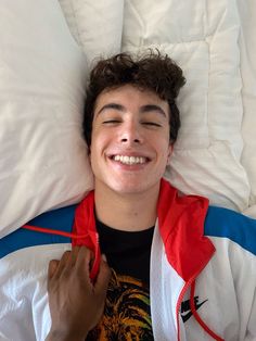 a young man laying in bed with his eyes closed and wearing a red white and blue jacket