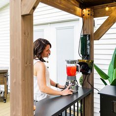 a woman standing on a porch with a blender in front of her and a potted plant behind her