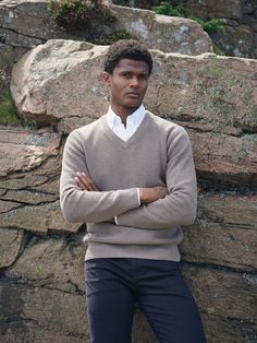 a man standing in front of a rock wall wearing a sweater and pants with his arms crossed