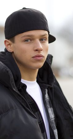 a young man in a black jacket and hat looking at the camera with a serious look on his face