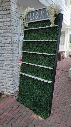 a sign that is covered in grass and flowers on the side of a building with brick walkway