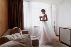 a woman standing in front of a window with a bouquet on her hand and wearing a wedding dress