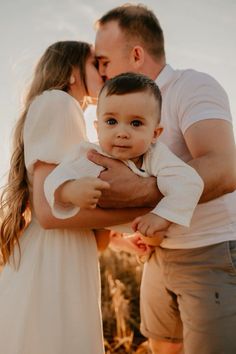 a man and woman holding a small child in their arms, while the baby is wearing a white dress