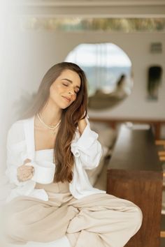 a woman sitting on the floor with her eyes closed and hands behind her head, wearing a white shirt and beige pants