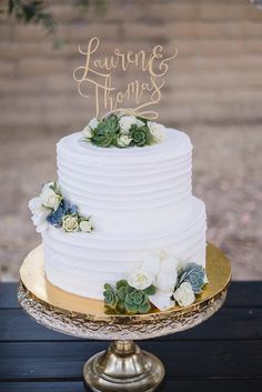 a white wedding cake with succulents and greenery sits on a gold platter