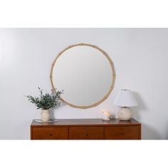 a round mirror sitting on top of a wooden dresser next to a white vase and lamp