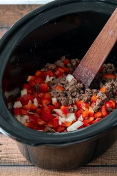 a slow cooker filled with ground beef and red bell peppers next to a wooden spoon