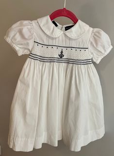 This blue and white Ralph Lauren dress is hand-smocked and the outer layer is made from delicately ribbed 100% white cotton. The smocking across the bodice features a darling anchor motif with white and blue striped  embroidery thread detailing. The voluminous sleeves elevate the overall look and have nice definition.  The garment is fastened using three (3) buttons up the back and a button belt that goes around the waist, (also fastened in the back). It's lined in feather-soft white cotton.  It White Smocked Short Sleeve Dress For Daywear, White Short Sleeve Smocked Dress For Daywear, White Short Sleeve Smocked Day Dress, White Smocked Dress With Short Sleeves For Daywear, Preppy Fitted White Dress, Classic White Smocked Dress For Spring, White Sailor Cotton Dress, Sailor Style White Cotton Dress, White Cotton Sailor Dress