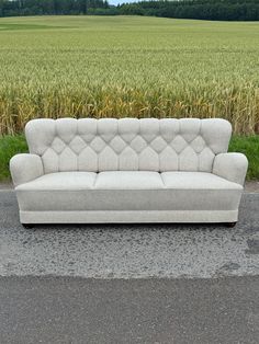 a white couch sitting on top of a cement road next to a corn field with trees in the background