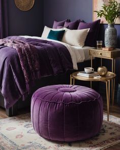 a bedroom with purple bedding, white pillows and a round foot stool in front of the bed