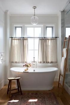 a white bath tub sitting under a window next to a wooden stool and shower curtain