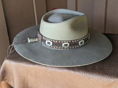 The Bandit is a stand out hat that captures the feeling of the American West. . The base of the hatband is distressed leather that is then layered with imprinted brown leather, bolo sliders and lots of nailspots. It is finished with this colorful feather combo. The brim of the LID custom hat body is 4" in diameter and the crown is 4.5". This is a stiff hat. Available in Putty or Pecan All hats are made to order. Allow up to 4 weeks for your hat to ship. Western Style Brown Hat With Flat Crown, Brown Fedora With Flat Crown For Ranch, Rustic Adjustable Felt Hat For Festival, Rustic Adjustable Felt Hat With Short Brim, Brown Fedora For Western-themed Events With Flat Crown, Adjustable Distressed Hats For Country Events, Brown Flat Crown Fedora For Western-themed Events, Distressed Adjustable Hats For Country Events, Brown Felt Hat For Western-themed Events