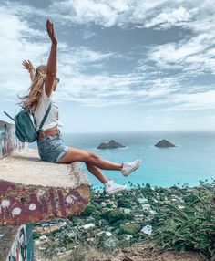 a woman sitting on the edge of a cliff with her arms up in the air