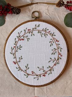 a white embroidery hoop with red berries and green leaves on it next to pine cones