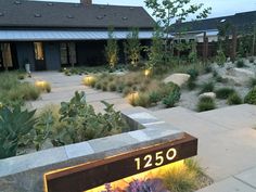 an illuminated sign in front of a house with plants and rocks on the ground next to it