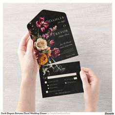 a person holding up a black and white wedding card with flowers on the front, in front of a wooden background